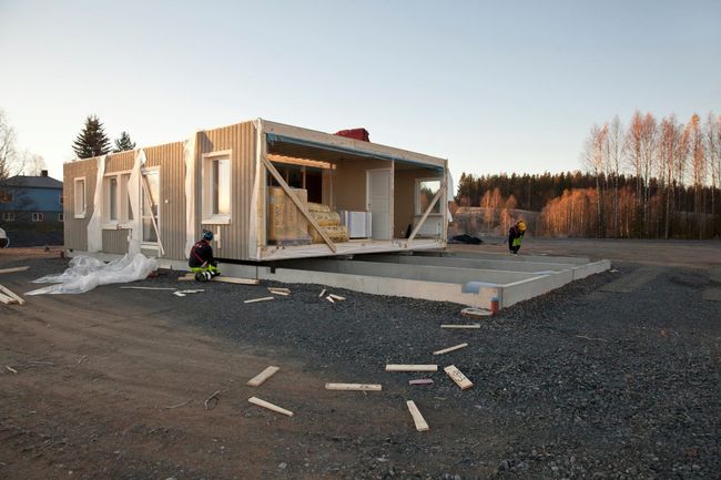 Två byggarbetare som står och arbetar brevid tre husmoduler för ett nyckelfärdigt grått hus.